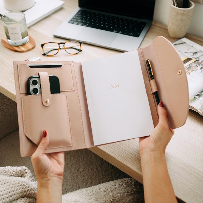 K&ouml;nnte beinhalten: Ein Portfolio aus rosa Leder mit einem Stifthalter, einer Tasche f&uuml;r ein Telefon und einer Tasche f&uuml;r einen Notizblock. Das Portfolio ist ge&ouml;ffnet und der Notizblock hat die Initialen "AM" auf der Vorderseite.
