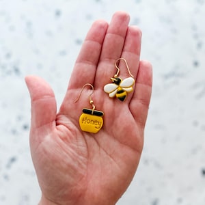 Puede incluir: Un par de pendientes dorados con diseños de abeja y tarro de miel. El pendiente de abeja tiene un esquema de color blanco, amarillo y negro. El pendiente de tarro de miel es amarillo con la palabra "Honey" y una parte superior negra. Los pendientes se sostienen en una mano.