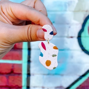 Pode incluir: Um par de brincos pendurados abstratos. Os brincos são brancos com pequenas manchas pretas e formas coloridas em vermelho, azul, verde e laranja. Os brincos são presos por um gancho dourado.