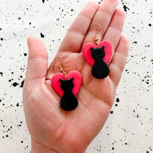 May include: A pair of handmade earrings featuring a black cat silhouette on a bright pink heart. The earrings have gold-colored hooks. The background is white with black paint splatters. These earrings are a fun accessory.