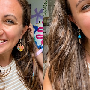 May include: A woman wearing a pair of earrings. One earring is a brown and white paint palette with colorful paint dots. The other earring is a blue and gold ice cream cone.