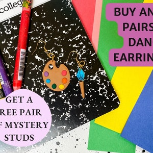 May include: A pair of dangle earrings featuring a paintbrush and a paint palette. The earrings are on a black and white notebook with colored pencils and the text "BUY ANY 2 PAIRS OF DANGLE EARRINGS GET A FREE PAIR OF MYSTERY STUDS".