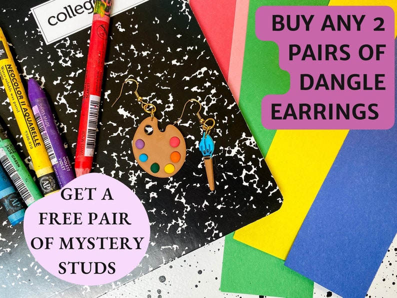 May include: A pair of dangle earrings featuring a paintbrush and a paint palette. The earrings are on a black and white notebook with colored pencils and the text "BUY ANY 2 PAIRS OF DANGLE EARRINGS GET A FREE PAIR OF MYSTERY STUDS".