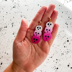 Op de afbeelding: Een paar roze en witte ombre schedel oorhangers met zwarte ster details. De oorbellen zijn gemaakt van polymer clay en hebben gouden oorhaken.