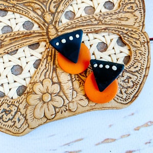 May include: A pair of black and orange geometric earrings with white dots. The earrings are shaped like a triangle with a rounded edge and a circle.