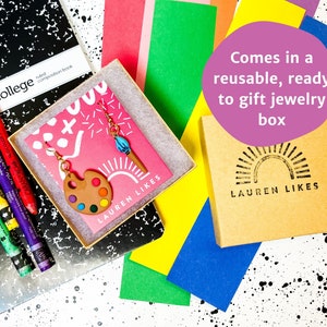 May include: A pair of colorful paint palette earrings with a paintbrush charm, packaged in a brown box with the text "LAUREN LIKES" and a sun graphic. The box is on a black and white background with colorful paper.