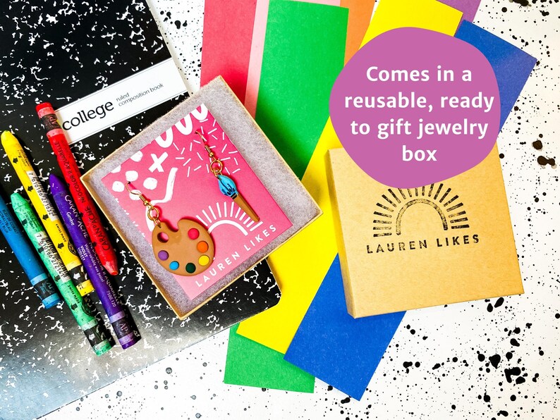 May include: A pair of colorful paint palette earrings with a paintbrush charm, packaged in a brown box with the text "LAUREN LIKES" and a sun graphic. The box is on a black and white background with colorful paper.
