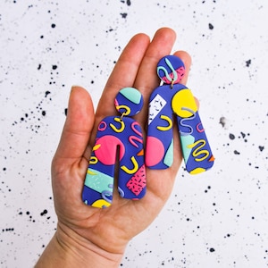 Könnte beinhalten: Ein Paar bunte geometrische Musterohrringe. Die Ohrringe sind aus Polymer Clay gefertigt und haben ein blaues, rosa, gelbes und grünes Farbschema. Die Ohrringe haben die Form eines abgerundeten Rechtecks mit einer abgerundeten Oberseite.