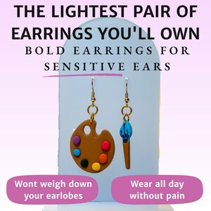 May include: A pair of lightweight earrings featuring a brown paint palette and a blue paintbrush charm. The earrings are advertised as being comfortable for sensitive ears. The text "The lightest pair of earrings you'll own" and "Bold earrings for sensitive ears" is displayed above the earrings. The text "Wont weigh down your earlobes" and "Wear all day without pain" is displayed below the earrings.