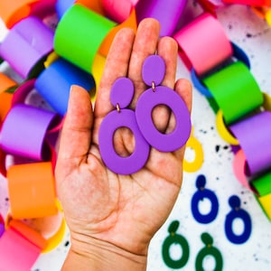 Könnte beinhalten: Ein Paar violette Statement-Ohrringe mit einem großen Kreisdesign. Die Ohrringe sind aus einem leichten Material gefertigt und haben eine goldfarbene Oberfläche.
