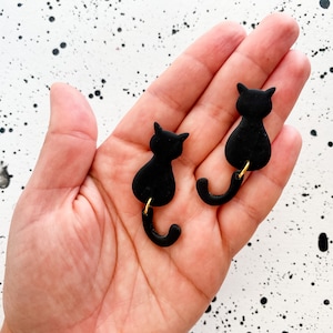 以下が含まれることがあります： 金色のアクセントが付いた黒い猫の形をしたイヤリングのペア。イヤリングはぶら下がっており、猫の尻尾が垂れ下がっています。