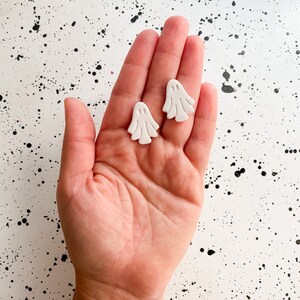 Puede incluir: Dos pendientes blancos en forma de fantasma con un diseño de cara simple. Los pendientes se muestran en una mano sobre un fondo blanco con motas negras.