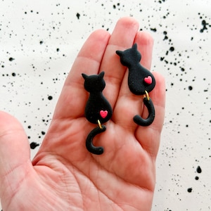 May include: Black cat earrings with a pink heart detail. The earrings are shaped like cats with curved tails and gold-colored hooks. The earrings are handmade and are a fun accessory.