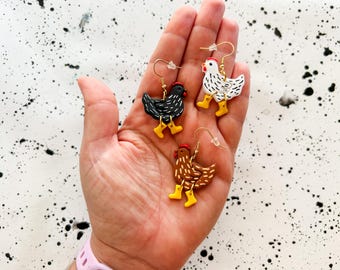 May include: Three pairs of handmade polymer clay earrings featuring chickens wearing yellow rain boots. The chickens are black and white, brown and white, and white.