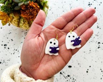 May include: A pair of handmade ghost earrings. Each white ghost holds a purple book and has black dot eyes. The earrings have gold-colored hooks. The earrings are held in a hand.