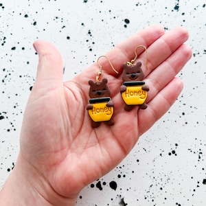 May include: A pair of handmade earrings featuring a brown bear holding a yellow honey pot. The honey pot has the word "Honey" printed on it. The earrings are on gold-colored hooks.
