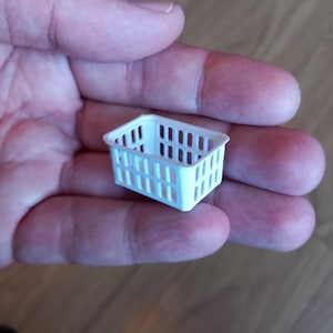 May include: A miniature white plastic basket, rectangular in shape with vertical slots. The small basket is held in a hand, highlighting its size and design. It's likely intended for decorative or craft use, with a simple, clean aesthetic.
