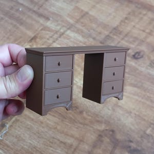May include: A miniature brown desk with six drawers and a rectangular top. The desk has a classic design with decorative drawer pulls and curved feet. The desk is held by a hand.