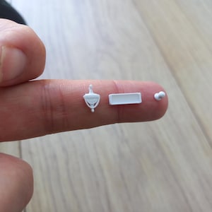 Peut inclure: Trois composants miniatures en plastique blanc comprenant un petit heurtoir de porte, un cadre rectangulaire et un minuscule bouton circulaire. Ces petits objets sont posés sur un doigt pour montrer leur échelle extrêmement réduite.