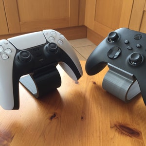 May include: A white and black PlayStation 5 controller and a black Xbox controller, each on a stand. The controllers are displayed on a wooden surface, highlighting their design and colour contrast.