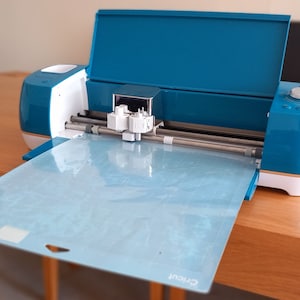 May include: A teal Cricut cutting machine with the lid open and a light blue cutting mat in place. The machine has a white base and a silver cutting blade.