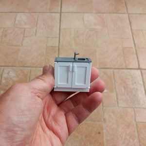 Könnte beinhalten: Ein kleiner weißer Küchenunterschrank im Miniaturformat mit zwei Türen und einer grauen Arbeitsplatte mit einem kleinen silbernen Wasserhahn. Der Schrank ist etwa 10 cm hoch.