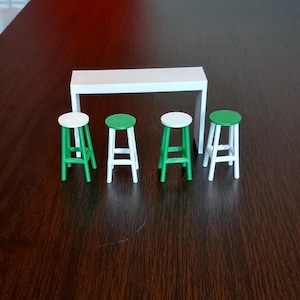 May include: A set of four miniature white and green stools with a white table top. The stools and table are made of plastic and are approximately 10 cm tall.