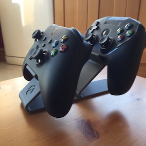 May include: Two black video game controllers are displayed on a black stand. The stand has a logo on the front. The controllers are on a wooden surface.