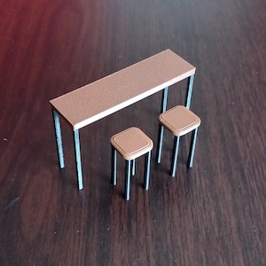 May include: A miniature brown table and two stools. The table has a rectangular top and black legs. The stools have square seats and black legs. The set is designed for a doll's house or similar small-scale setting.