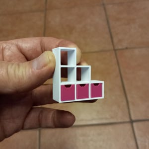 May include: A miniature white stepped shelf unit with three pink drawers. The unit has an open design with a stepped configuration, perfect for displaying small items. The drawers are a vibrant pink.