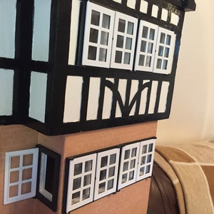 May include: A miniature model of a house with black and white timber framing and white windows. The house has a brown brick facade and a white roof.