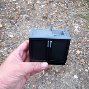 May include: A miniature black cabinet with two doors and a grey countertop with a small sink and faucet.