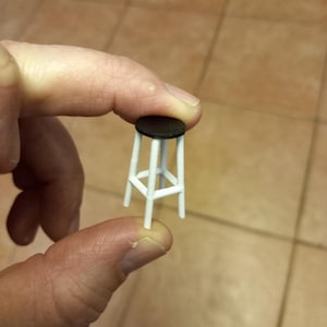 May include: A miniature white stool with a dark brown seat is held between two fingers. The stool has four legs and a circular seat. The legs are connected by horizontal supports.