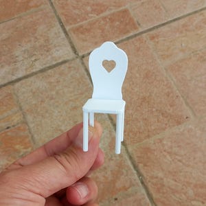 May include: A miniature white chair featuring a heart-shaped design on the backrest. The chair has a square seat and four straight legs. The chair is held in a hand, highlighting its small size, likely a dollhouse chair.