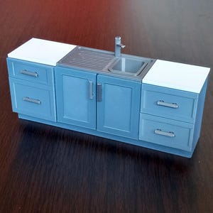 May include: Miniature blue kitchen cabinet set with a stainless steel sink, tap, and countertop. The set includes drawers and cabinet doors with silver handles. The countertop is white.