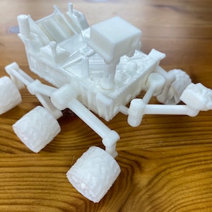May include: A white 3D printed model of a Mars rover with six wheels and a robotic arm.
