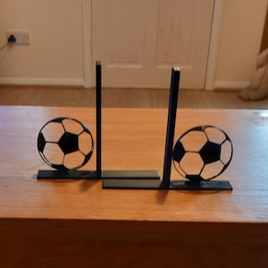 May include: A decorative soccer-themed display featuring two black and white soccer balls. The balls are held in place by a black metal frame, creating a goalpost design. The display sits on a wooden surface.