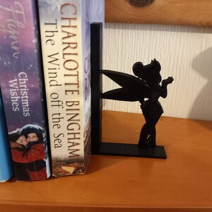 May include: Black bookend featuring a silhouette of Tinkerbell, a fairy with wings, holding a wand. The bookend is on a wooden shelf with books behind it.