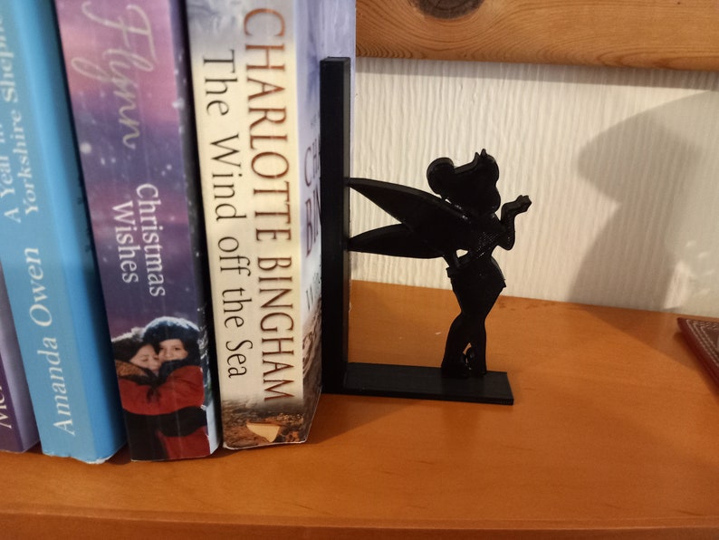 May include: Black bookend featuring a silhouette of Tinkerbell, a fairy with wings, holding a wand. The bookend is on a wooden shelf with books behind it.