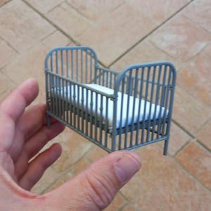 May include: A small, grey metal cot with vertical bars and a white mattress. The cot is held in a hand, highlighting its miniature scale. The design is simple and classic, suitable for a dollhouse or decorative display.