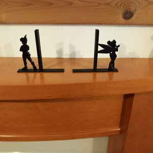 May include: A pair of black metal bookends featuring silhouettes of Peter Pan and Tinkerbell. The bookends are on a wooden surface.