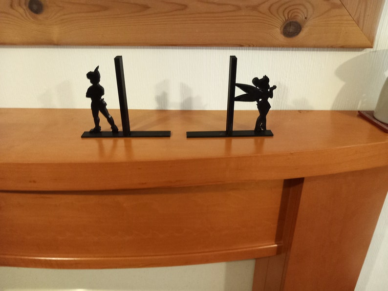May include: A pair of black metal bookends featuring silhouettes of Peter Pan and Tinkerbell. The bookends are on a wooden surface.