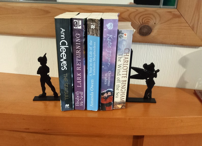 May include: Black metal bookends featuring a silhouette of Peter Pan and Tinkerbell. The bookends are holding up a stack of books with titles like "The Seagull", "Lark Returning", "A Year in the Life of the Yorkshire Shepherdess", "Christmas Wishes", and "The Wind off the Sea".