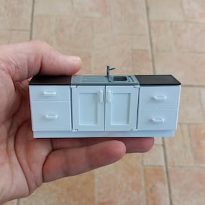 May include: A miniature white kitchen sink with two cabinets on either side and a black countertop. The sink has a small faucet and a drain.