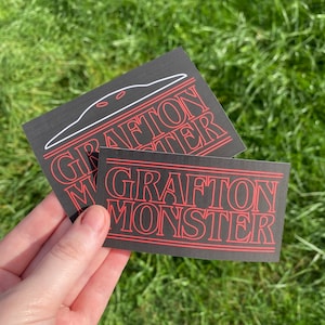 May include: Two black rectangular stickers with the text "GRAFTON MONSTER" in red, stylized font. One sticker has a white outline of a creature above the text. The stickers are held against a green grassy background.