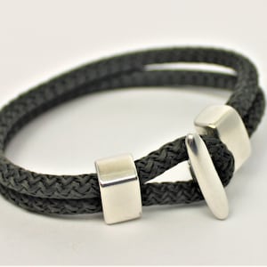 Könnte beinhalten: Ein schwarzes geflochtenes Armband aus Seil mit silbernen Metallakzenten. Das Armband hat einen Schiebeverschluss.
