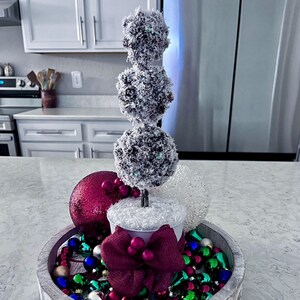 May include: A white wooden tray with a white metal pot and a three-tiered Christmas tree made of white and brown balls. The tray is decorated with a red bow, red and green baubles, and a white fluffy garland.