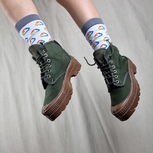 May include: A pair of dark green canvas boots with brown rubber soles. The boots are laced up and have a casual, comfortable look. The person wearing the boots is also wearing white socks with a rainbow cloud pattern.