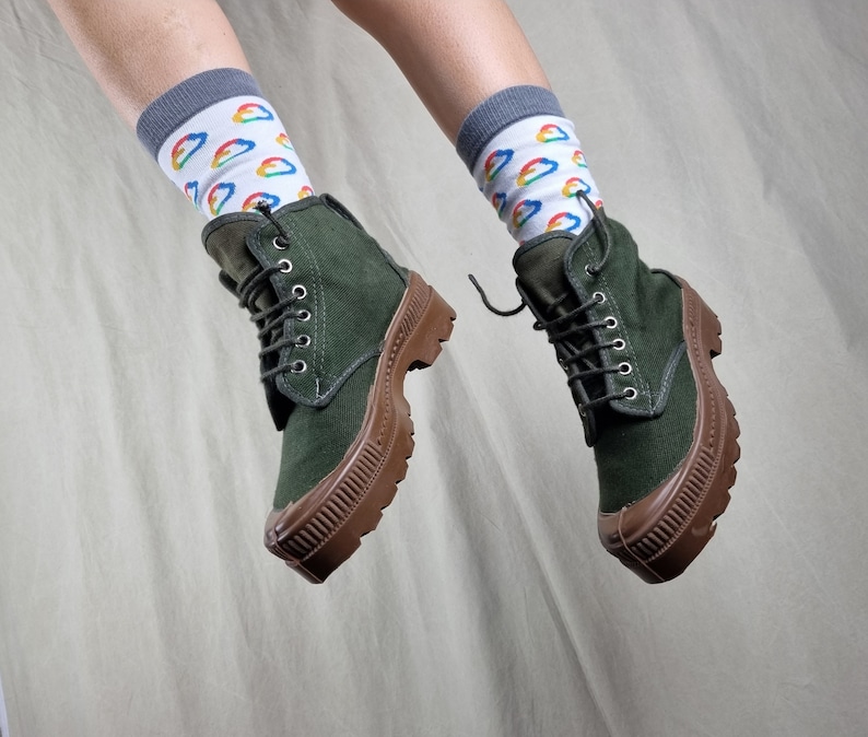May include: A pair of dark green canvas boots with brown rubber soles. The boots are laced up and have a casual, comfortable look. The person wearing the boots is also wearing white socks with a rainbow cloud pattern.