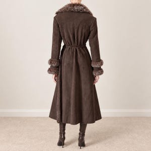May include: A long, dark brown coat with a suede-like texture, featuring a wide faux fur collar and matching faux fur trim on the cuffs. The coat has a fitted waist with a tie belt and a flared skirt, paired with dark brown knee-high boots.